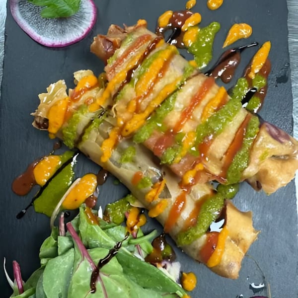 Crispy spring rolls drizzled with green, orange, and brown sauces, served with a side salad.