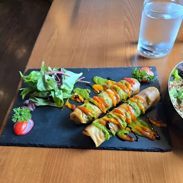Two spring rolls with green and orange sauce, salad, and a glass of water on a wooden table.