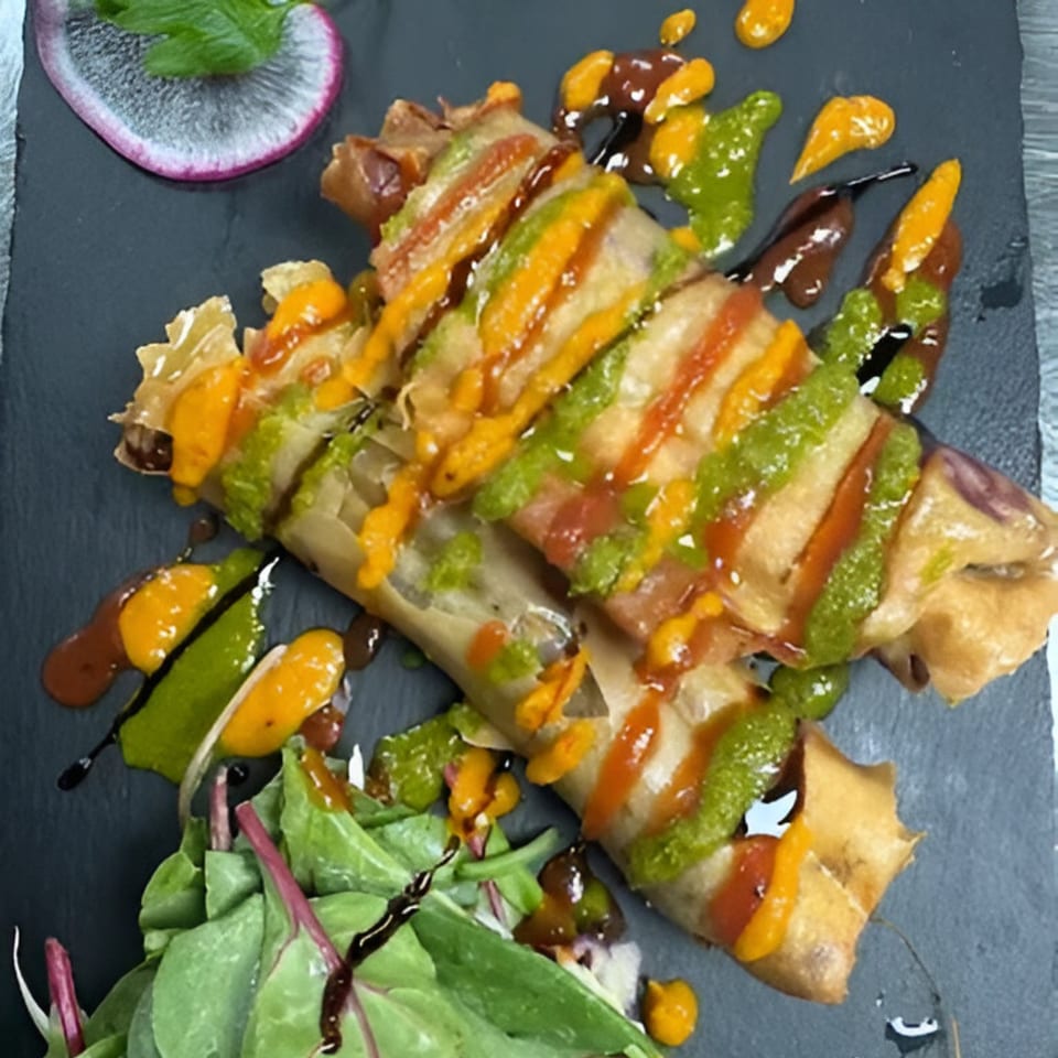 Crispy spring rolls drizzled with colorful sauces, served with a side salad on a dark plate.